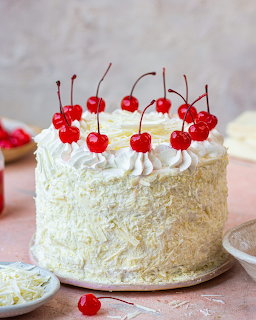 White frosted layer cake decorated with whipped cream, white chocolate shavings, and candied cherries on stems