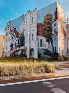 Large photorealistic street art mural on a building exterior showing historic town architecture with a bridge and trees.