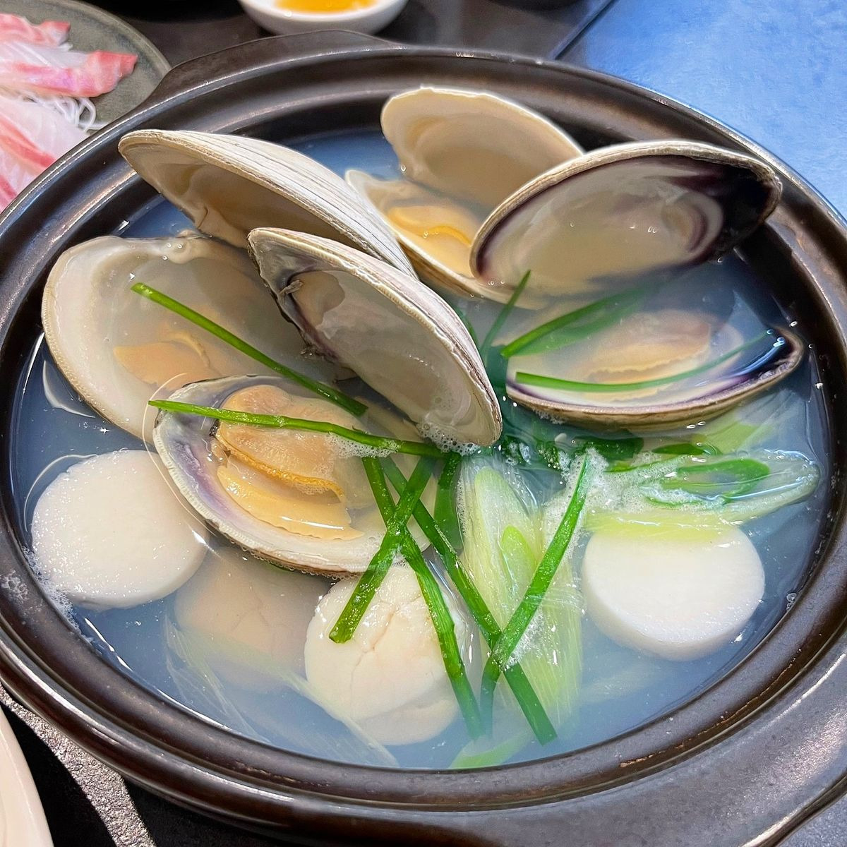 Bowl of clear clam soup with open clam shells, white fish balls, and green onion garnish