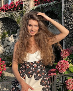 Smiling woman with long wavy brown hair posing in a flower garden with pink bloms