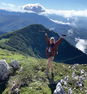 Woman hiker celebrating on a mountain summit with arms raised holding trekking poles against green mountain backdrop