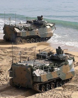 Two camouflaged AAVP 7A1 amphibious assault vehicles parked on a sandy beach near the ocean