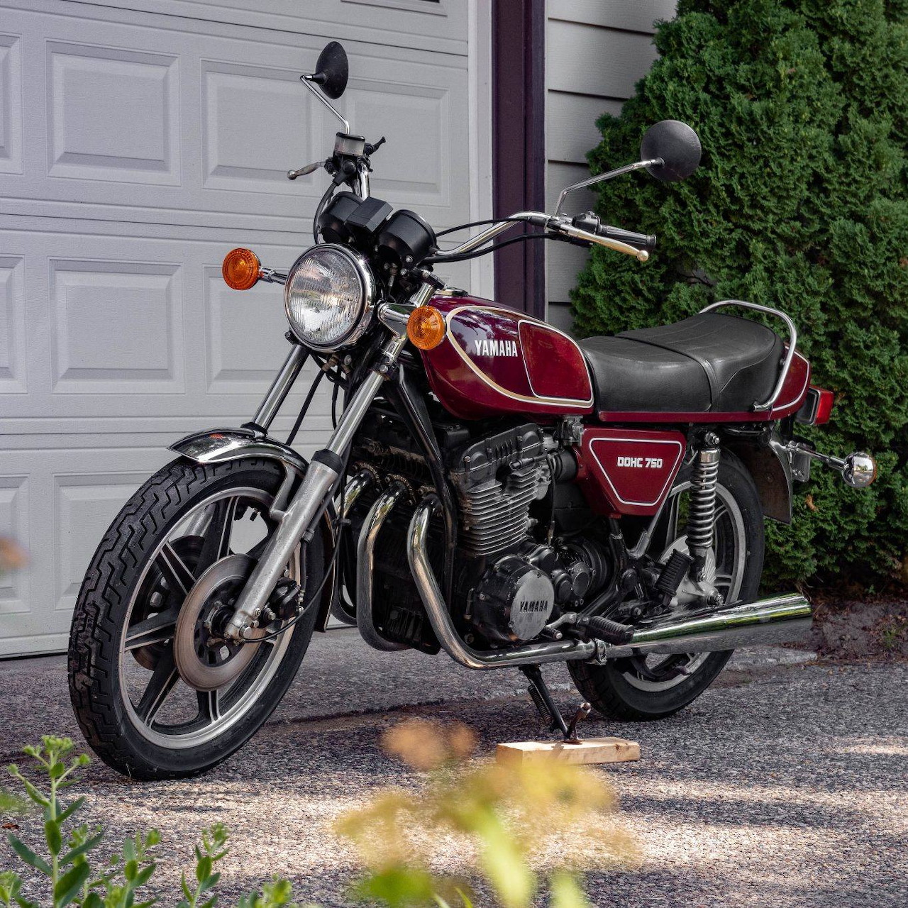 Burgundy 1977 Yamaha XS750 motorcycle parked in front of white garage door with green bushes in background