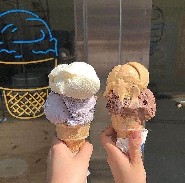 Two hands holding ice cream cones with white and peach scoops in front of neon ice cream sign