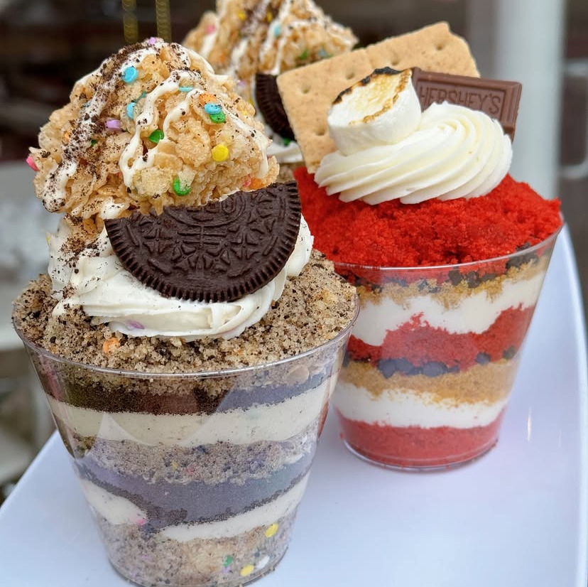 Two layered dessert parfaits with cookies, whipped cream, and colorful cake layers in clear plastic cups