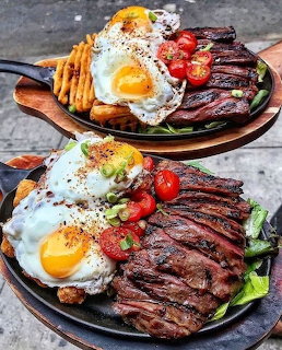 Two black plates with steak, fried eggs, fries, and tomatoes on marble counter