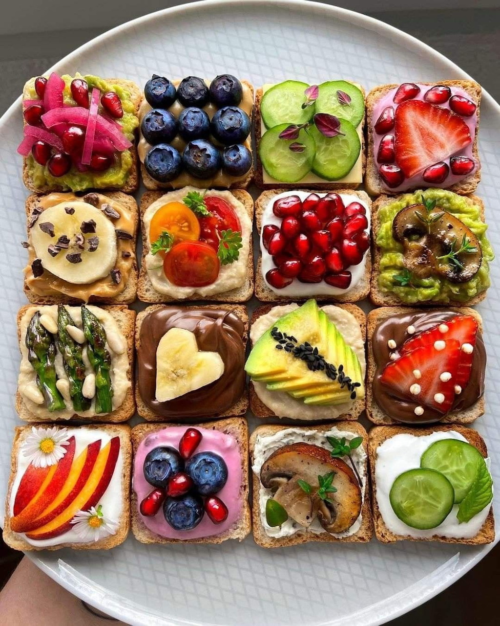 16 decorated toasts on a white plate with colorful fruit and vegetable toppings