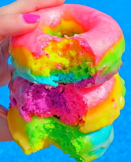 Hand holding a stack of four rainbow-glazed donuts with colorful icing against a blue background