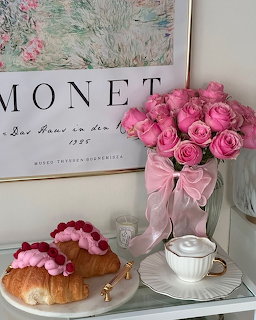 Vase of pink roses with ribbon, raspberry croissant on plate, white coffee cup, and Monet print in background