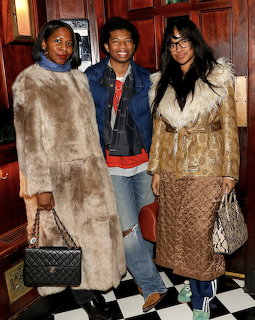 Three people posing at a Vogue party, two in fur coats and one in a denim jacket, indoors.