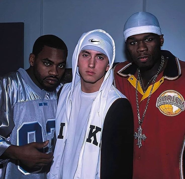 Three hip hop artists posing indoors, with Eminem centered in a white hood and durag