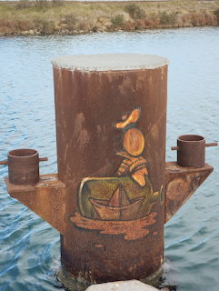 Rusty canal moring bollard with painted folk art of a person rowing a boat, water in background