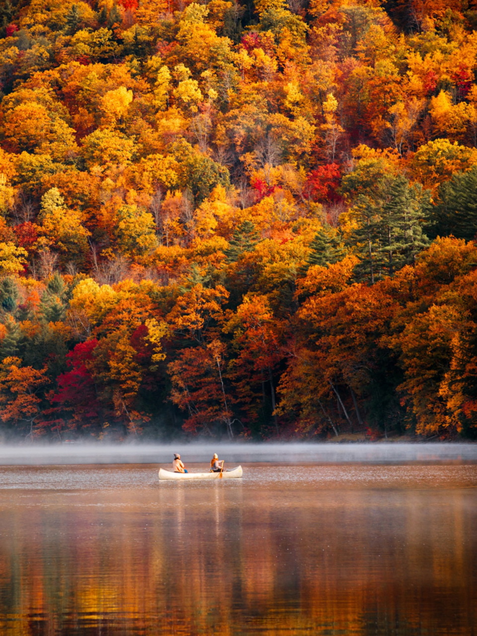 Two canoeists on a misty lake below a colorful autumn forest reflected in calm water.