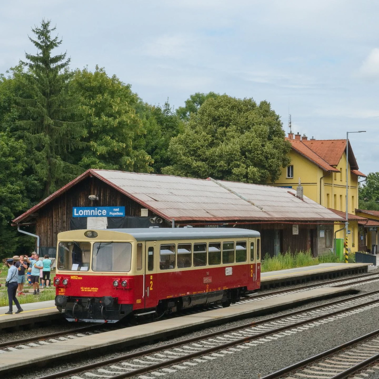 Red vintage railcar stopped at a rural train station platform with yellow building and trees in background