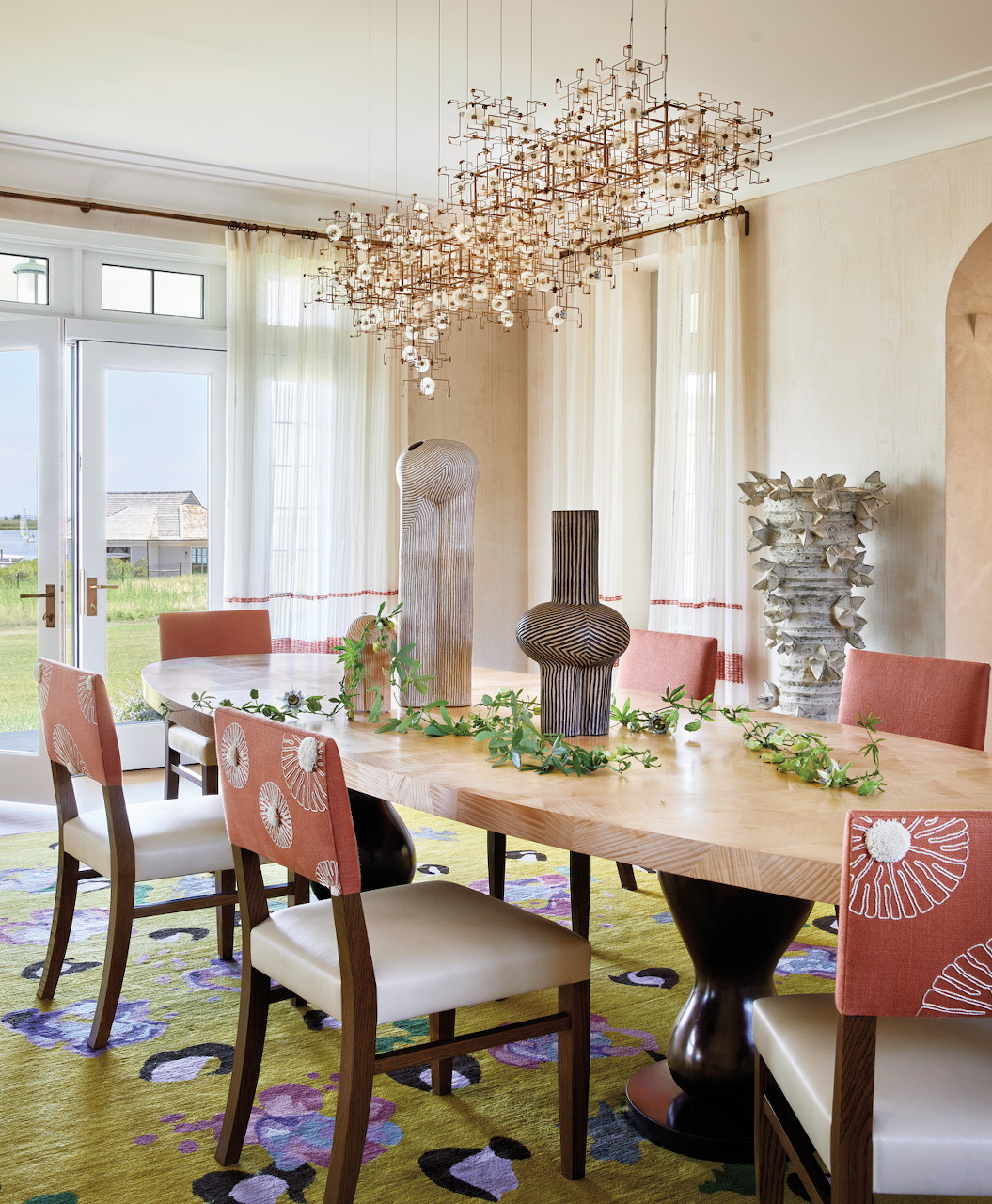 Dining room with wooden table, coral chairs, modern chandelier, and large windows