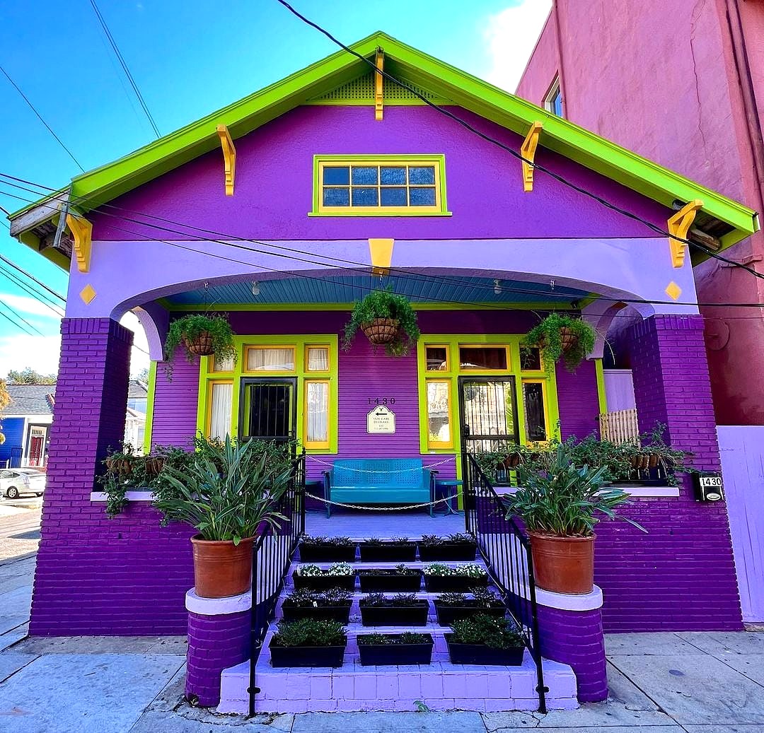 Bright purple house with green gabled roof, yellow trim and door, front porch with plants
