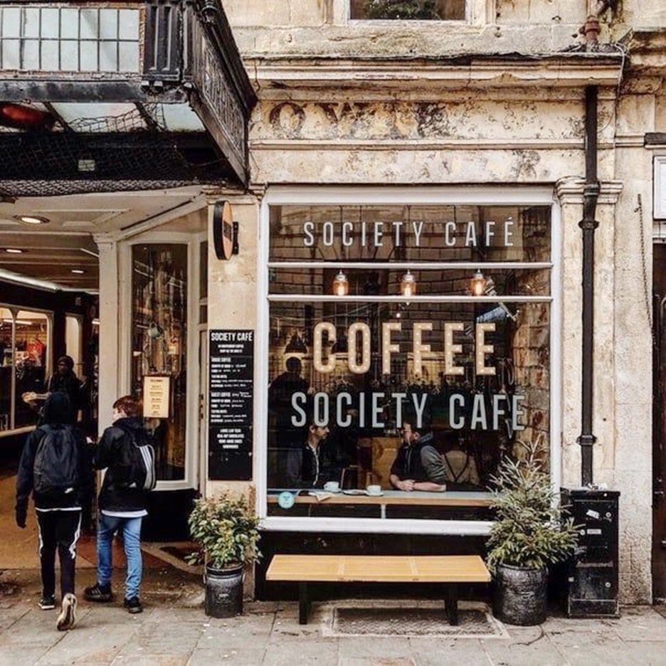 Stone facade of Society Cafe with large windows, outdoor bench, and two people walking by in Bath, England