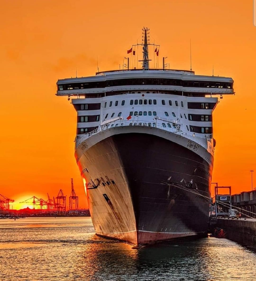 Large cruise ship Queen Mary 2 docked at port with a bright orange sunset sky behind it