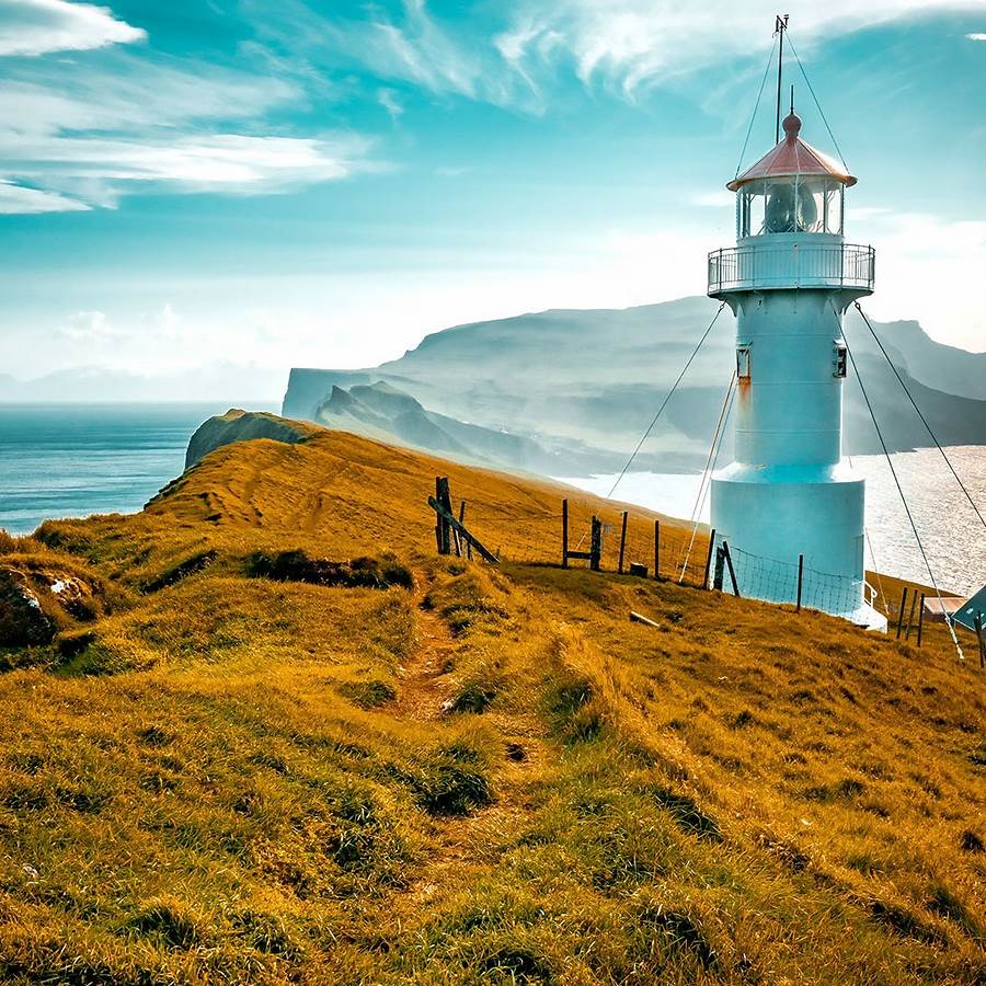 White lighthouse on grassy cliff with ocean and mountains under turquoise sky