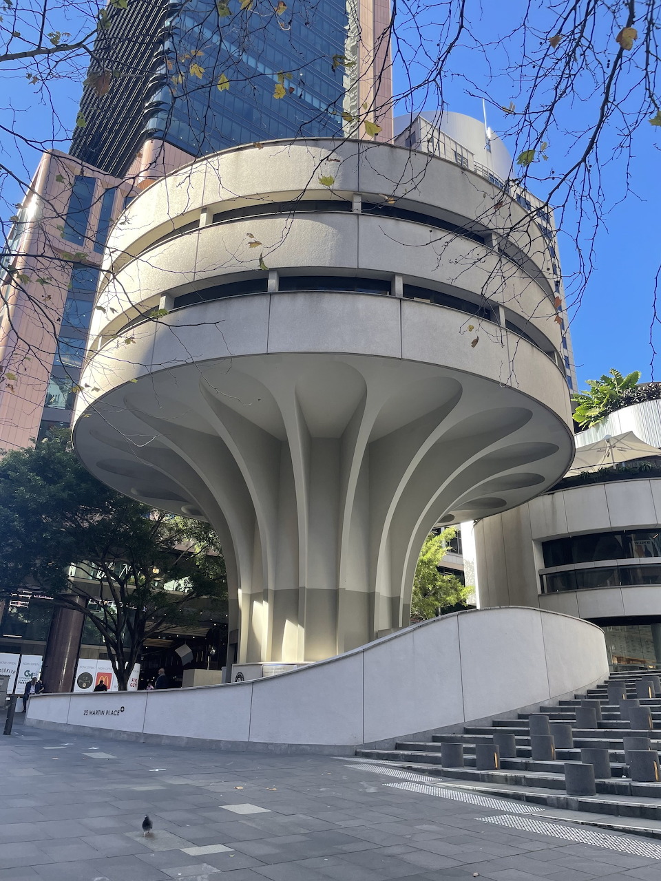 Circular brutalist building with flared white columns in an urban plaza with trees and skyscrapers