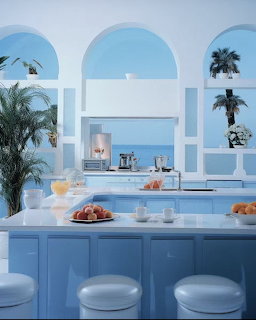 Pastel blue coastal kitchen with stools, fruit, palm leaves, and an ocean view through arched openings