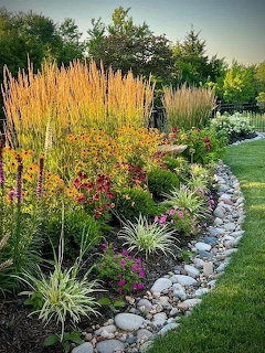 Curved garden border with colorful flowers, ornamental grasses, and white stone edging beside lawn