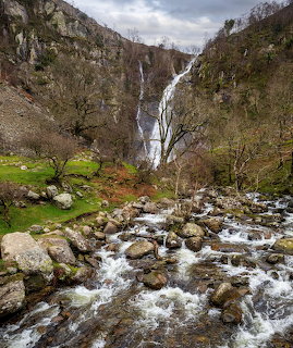 Aber Falls waterfall in Snowdonia, Wales, with a rocky stream flowing in the foreground and bare trees on the hillside.
