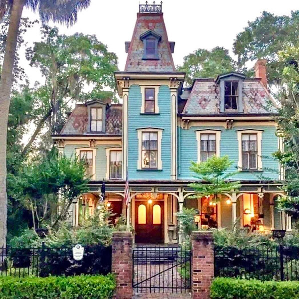 Victorian house with turquoise facade, decorative tower, and warm lit windows at twilight behind wrought iron gate