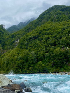 Turquoise river with white rapids flowing through a valley of green forested mountains under cloudy skies