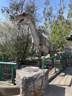 Large dinosaur statue beside a fenced walkway in an outdoor park with trees and rocks