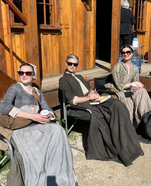 Three women in period dresses seated outside a wooden wooden building in sunlight