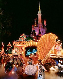 Disney castle lit up at night with illuminated parade floats in foreground