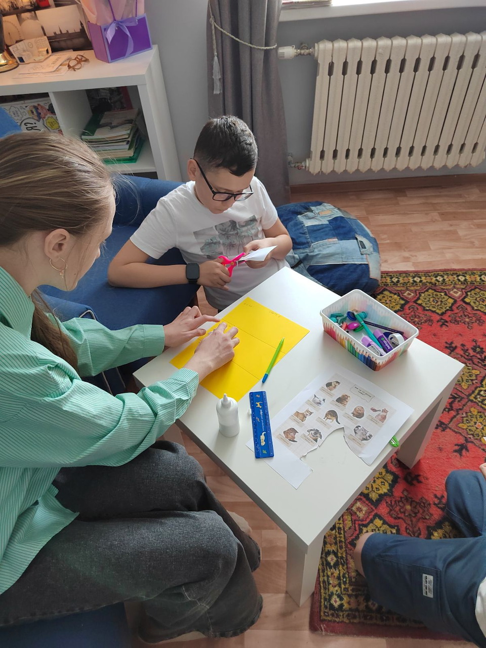 Two kids crafting at a white table with yellow paper, scissors, and small decorations indoors