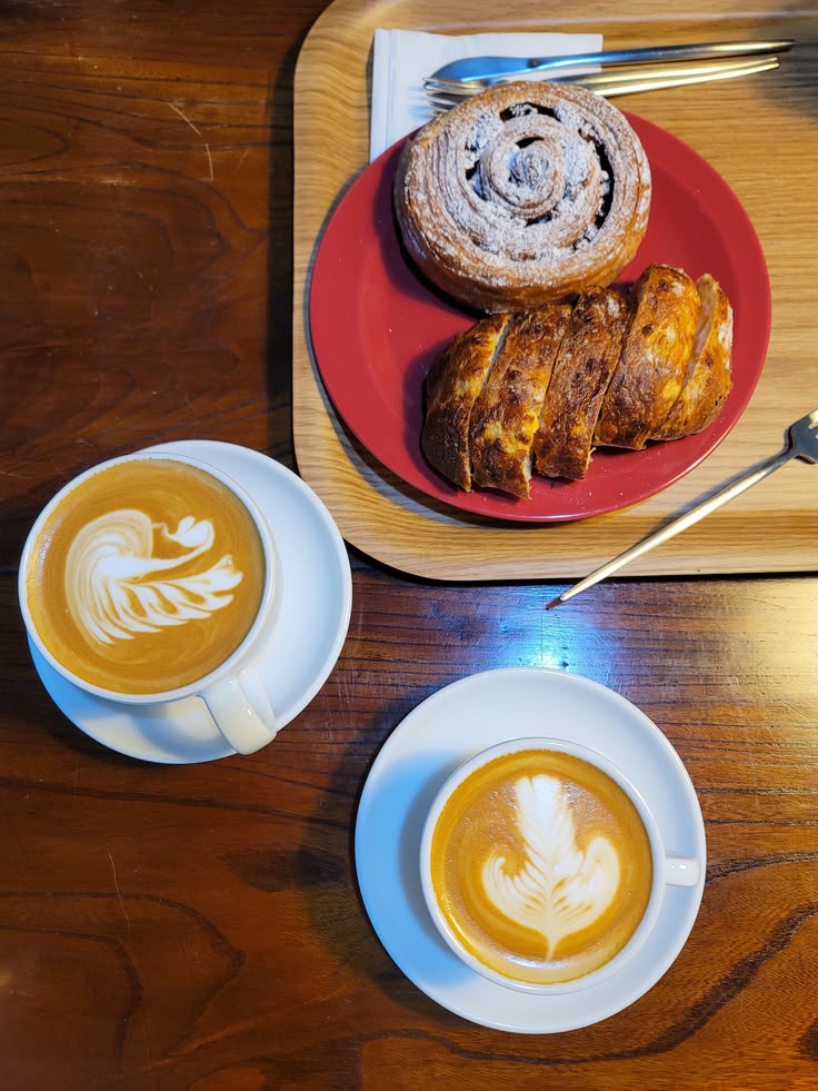 Overhead view of two lattes with latte art, a croissant, and a cinnamon roll on a red plate and wooden tray