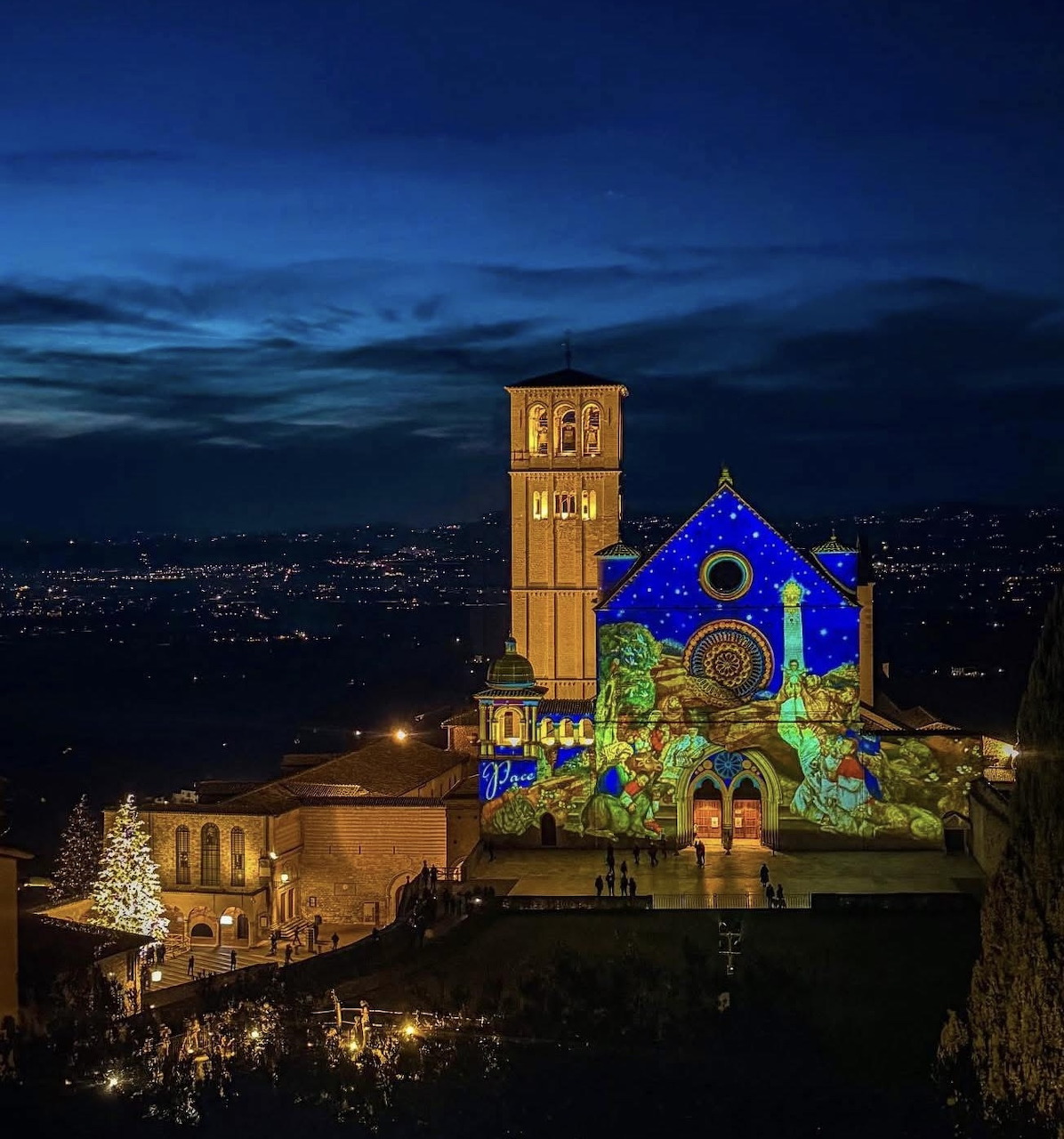 Assisi basilica illuminated at night with blue Christmas lights and decorated trees against twilight sky