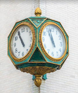 Ornate green and gold street clock with two white faces showing 10:56, mounted on pole against white building
