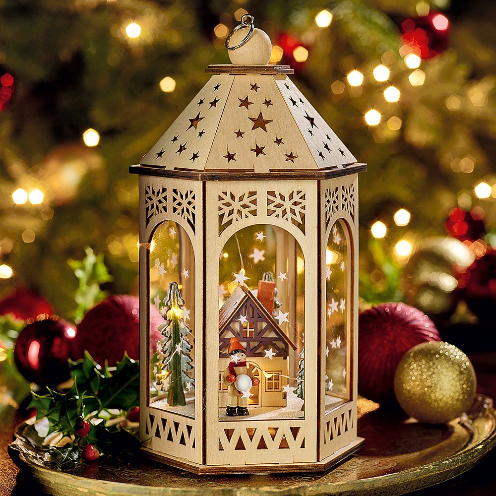 Illuminated white Christmas lantern with ornate cutouts on golden tray with holiday decorations and blurred lights