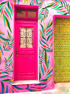 Bright pink wooden door framed by colorful painted tropical leaves on white wall with yellow shuttered window
