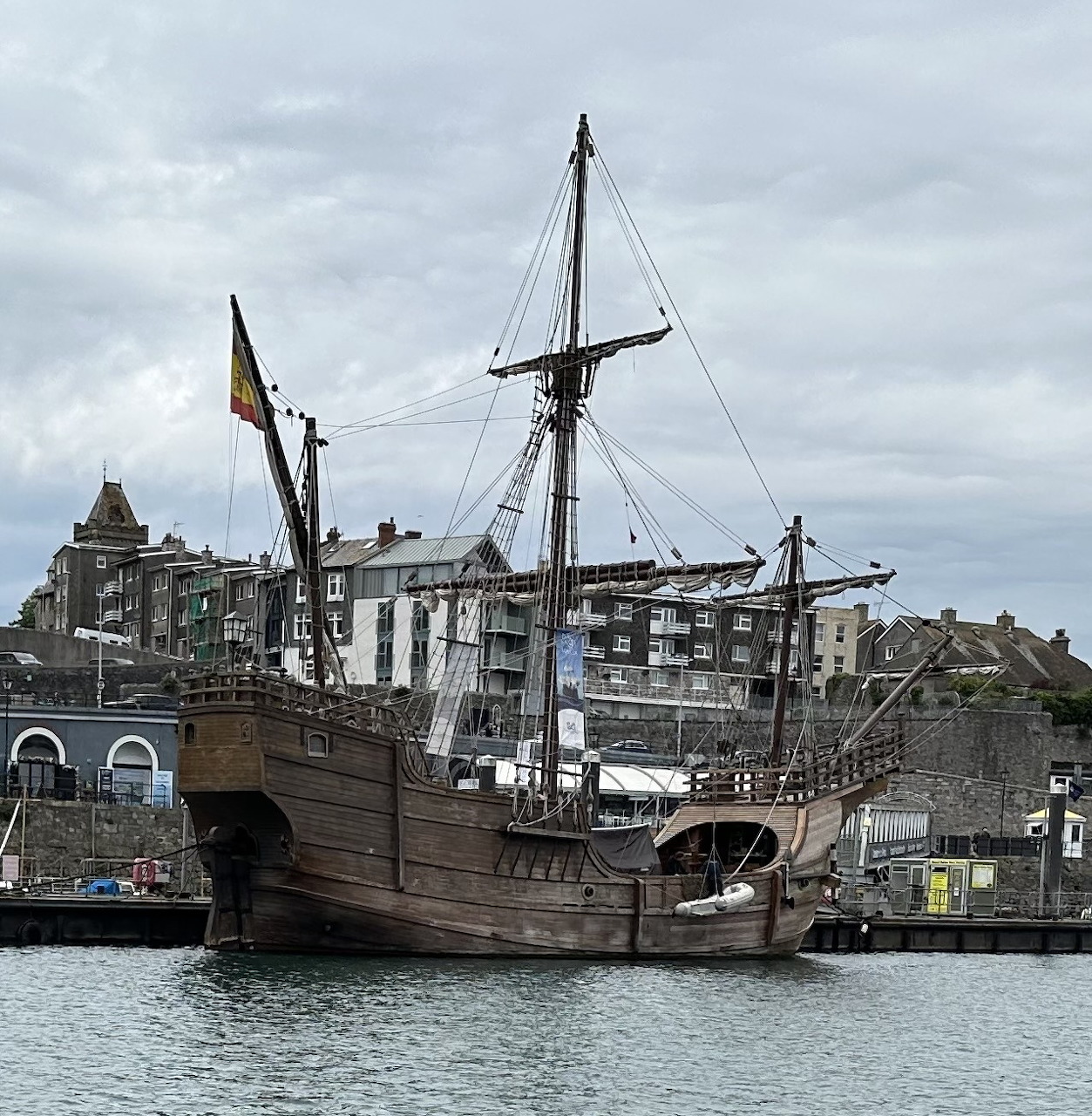 Historic wooden sailing ship docked in Plymouth harbor with waterfront buildings behind it