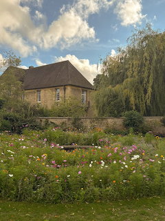 Stone cottage behind a wildflower garden with pink and white cosmos under a partly cloudy sky