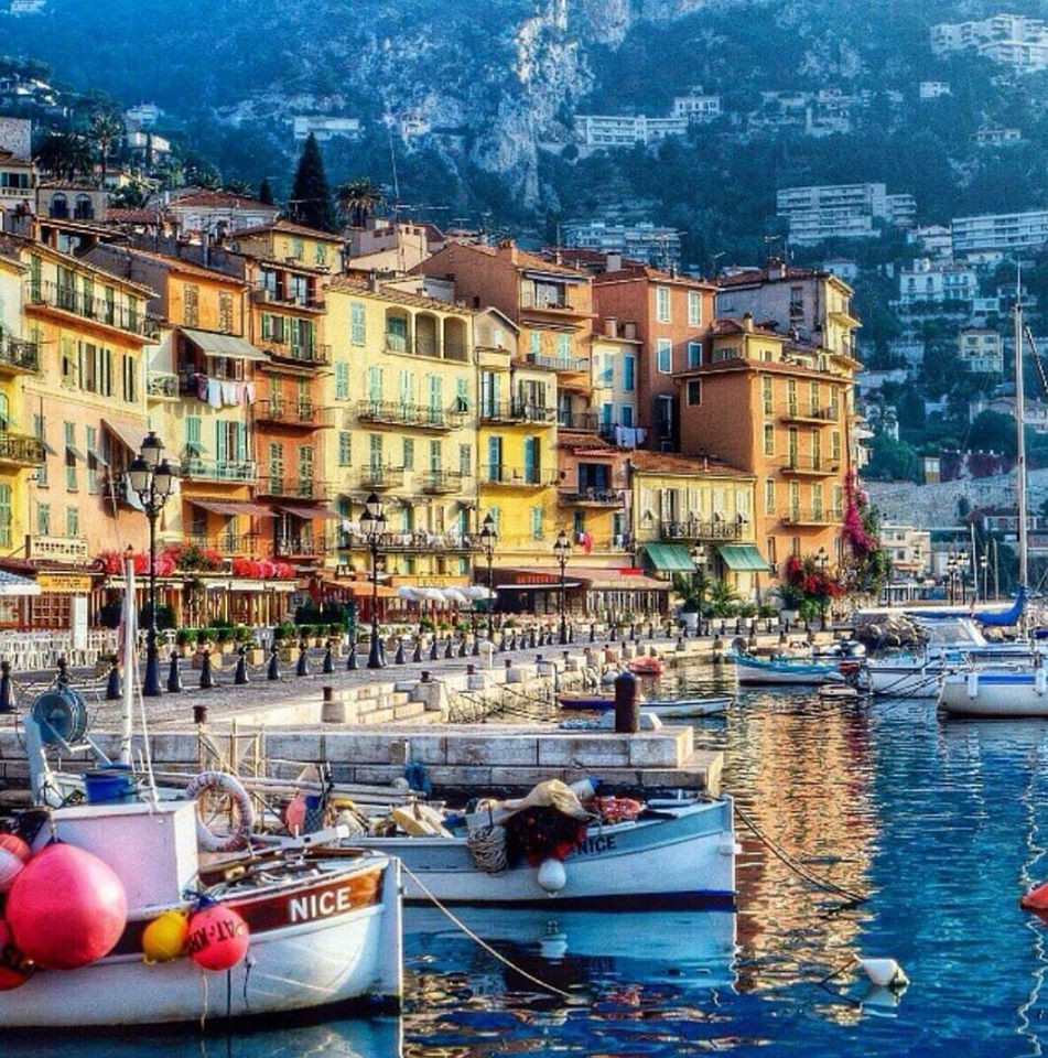 Harbor scene in Nice, France with colorful buildings, boats, and hillside backdrop at twilight