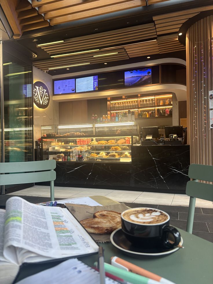 Latte with latte art on a cafe table with an open book and pastry, modern coffee bar behind.