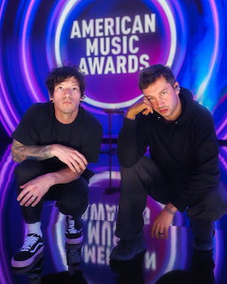 Two men crouching and posing in front of a neon purple and blue American Music Awards backdrop