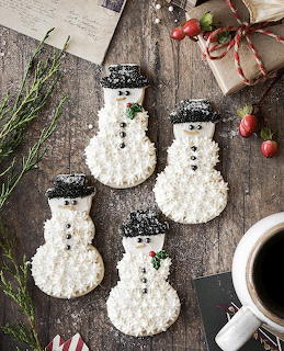 Four snowman-shaped cookies covered in white coconut with black hats, arranged on weathered wood with Christmas decorations