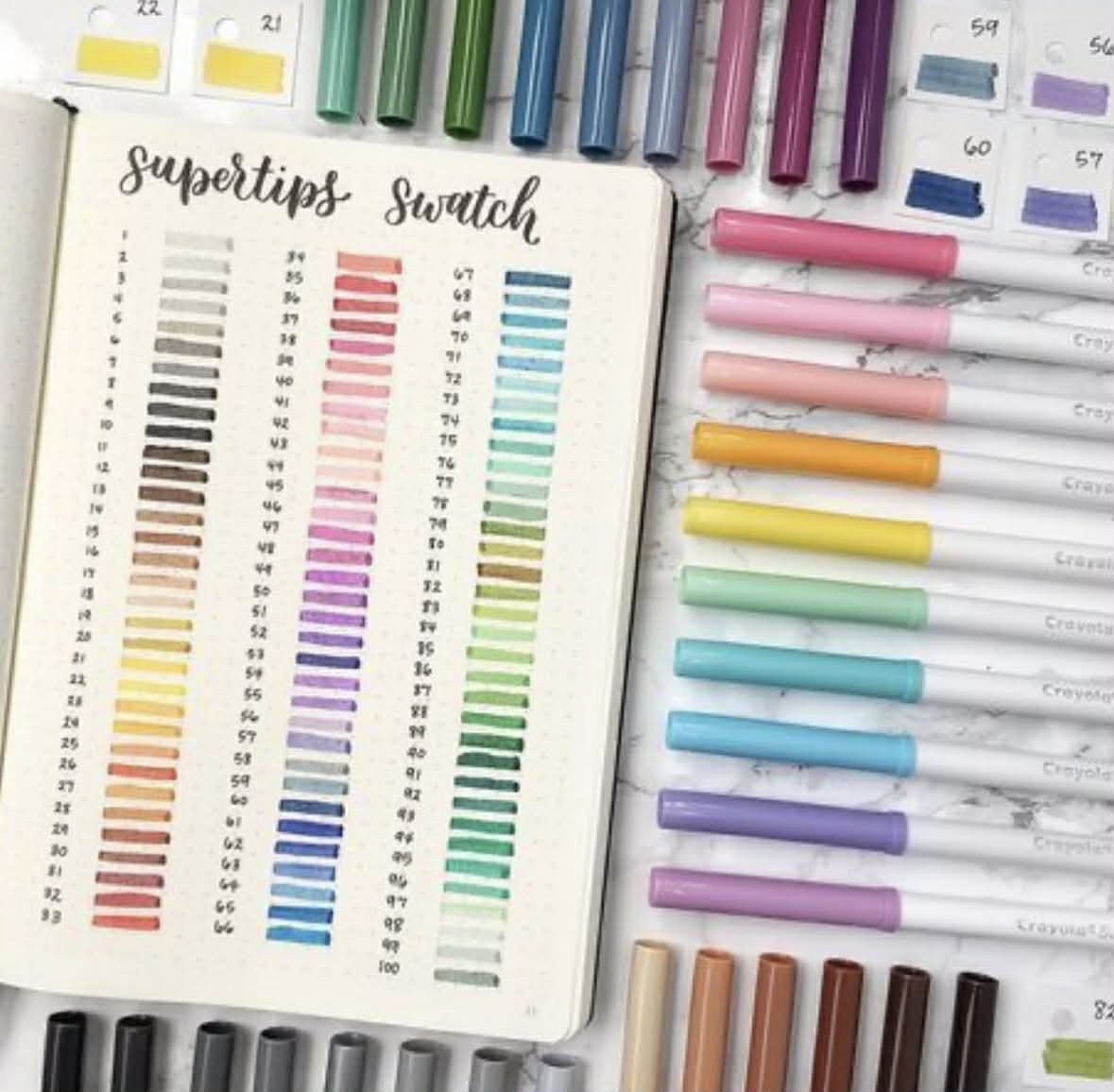 Notebook with color swatch chart surrounded by rainbow markers on marble background