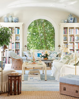 Bright living room with white sofas, bookshelves, wicker chairs, and a large arched window with greenery