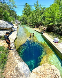 Natural stream with deep blue swimming hole, rocks, and green trees with people swimming
