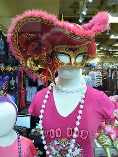 Mannequin wearing ornate pink and gold Mardi Gras mask with feathers and rose, pink shirt, and bead necklaces