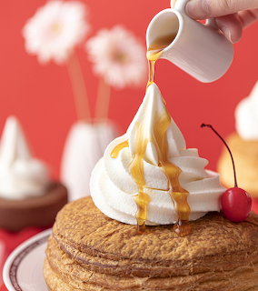 Stack of pancakes with whipped cream tower, syrup being poured, and cherry on top