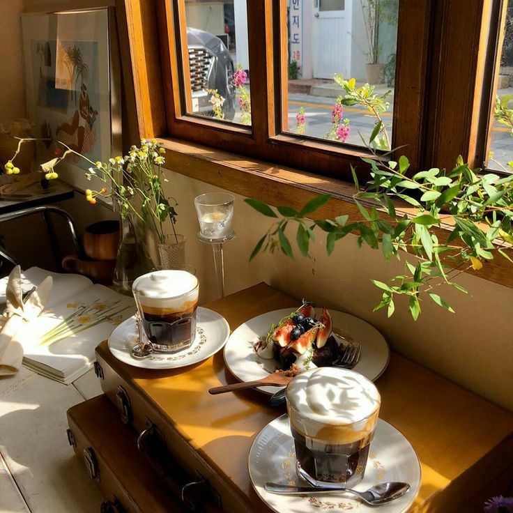 Layered coffee drinks and pastries on a sunlit cafe table beside a wooden window and plants